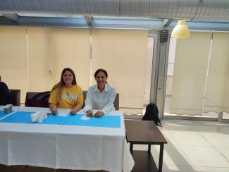 Beyaz örtü üzerine mavi runner bulunan masanın gerisinde iki kadın var. Solda sarı tişörtü, uzun açık kumral saçları olan kadın tebessüm ediyor. Sağdaki kadın beyaz bluzu, toplu saçlarıyla gülümsüyor.