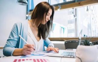 Genç bir kadın, modern bir ofis ortamında bir masada oturuyor. Önünde açık bir dizüstü bilgisayar var ve bir elinde kalem tutarak kâğıt üzerindeki grafiklere bakıyor. Üzerinde beyaz bir bluz ve açık mavi bir ceket var. Masanın üzerinde ayrıca bir bitki saksısı bulunuyor.