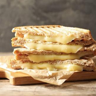 Ahşap bir kesme tahtası üzerinde üst üste dizilmiş üç adet tost dilimi. Tostların içinden erimiş peynir akıyor ve peynirin sıcaklığı nedeniyle hafifçe erimiş durumda. Tost ekmekleri ızgara izleri taşıyor ve altlarında kahverengi bir yağlı kâğıt var. Arka plan bulanık bir gri tonunda.