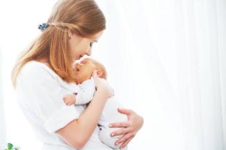 Bir anne ve kucağında tuttuğu bebek. Anne, bebeğini sevgiyle göğsüne yaslamış ve ona gülümsüyor. İkisi de beyaz giysiler giymiş. Arka plan çok aydınlık ve beyaz. Anne ve bebek arasında yakın ve sevgi dolu bir bağ hissediliyor.