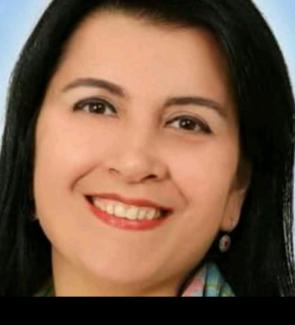 Düz, siyah saçları var ve saçları omuz hizasında. Ten rengi açık, dudaklarında kırmızı bir ruj var ve gülümsüyor. Yüz hatları belirgin, dişleri görünüyor. Bir kulağında küçük, yuvarlak küpe var. Boynunda renkli bir fular veya atkı göze çarpıyor. Arka plan açık mavi ve sade.