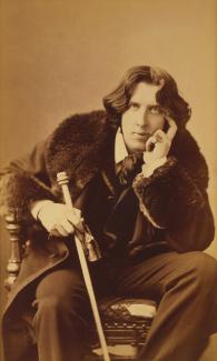 Oscar Wilde, sandalyede oturuyor. Üzerinde kalın kürklü bir palto var, içinde de gömlek ve kravatla oldukça resmi bir şekilde giyinmiş. Saçları orta uzunlukta, yanlara doğru dalgalı. Sol elini yüzüne dayamış ve düşünceli bir şekilde ileriye bakıyor, sağ eliyle bir değnek veya baston tutuyor.