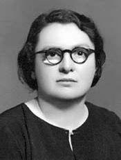 Siyah beyaz portre fotoğrafı.Mebrure Gönenç, yuvarlak çerçeveli gözlük takıyor. Kısa, koyu renkli saçları kulak hizasında dalgalı bir şekilde duruyor. Ciddiye yakın bir ifadeyle kameraya bakıyor. Üzerinde yakasız, koyu renkli bir üst var; kıyafetin ön kısmında bir fermuar veya dikkat çekici bir dikiş hattı bulunuyor. Arka plan düz ve açık 