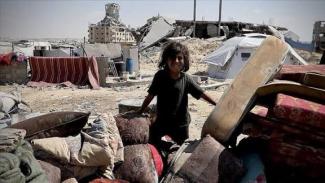 Enkazın ve çadırların önünde, yıpranmış kıyafetli küçük bir kız çocuğu. Arka planda yıkılmış binalar, çadırlar ve yere serilmiş battaniyeler var. Çocuğun yanında üst üste yığılmış minderler, döşekler dikkat çekiyor. Çocuğun ifadesi ciddi ve hüzünlü. Arka planda kısmen ayakta kalmış bir bina ve çadırlardan oluşan geçici barınma alanları görülüyor. Her şey tozlu ve düzensiz.