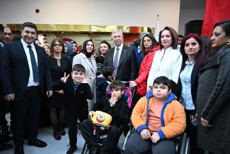 Kalabalık bir grup insan kapalı bir ortamda bir araya gelmiş. Grupta yetişkinler ve çocuklar var. Ön planda iki çocuk tekerlekli sandalyede oturuyor; ilki turuncu bir ceket giymiş, diğeri önünde Sünger bob desenli bir yastık tutuyor ve el sallıyor. Bir başka çocuk önde, siyah giysiler içinde, gülümseyerek zafer işareti yapıyor. Arkada takım elbiseli ve kravatlı bir adam ortada, çevresinde farklı yaş ve cinsiyetten insanlar var. Arka planda altın renginde bir tabela ve Türk bayrağı da sağ tarafta yer alıyor.