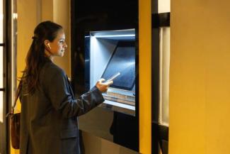 Banka ATM’sinde işlem yapan bir kadın. Kadın, koyu renk bir ceket giymiş, saçları arkadan toplanmış ve kulağında kablosuz kulaklık takılı. Bir elinde telefon tutuyor ve makinenin ekranına bakıyor. Makinanın çevresi siyah çerçeveli ve ortamda sıcak tonlu bir aydınlatma var.