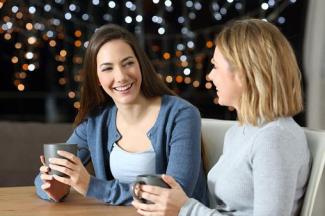 Bir kafede veya evde oturmuş, ellerinde kupa tutan iki kadın. İkisi de gülümsüyor ve birbirleriyle sohbet ediyorlar. Arka planda loş ışıklar ve bokeh efektiyle görünen küçük parlak noktalar var. Kadınlardan biri uzun kahverengi saçlı, diğeri kısa sarı saçlı.