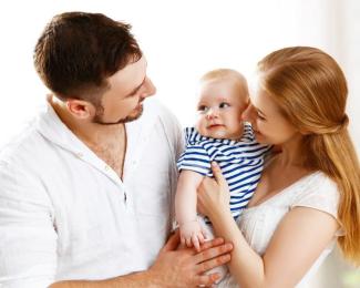 Anne, baba ve bebekten oluşan bir aile portresi. Anne ve baba, kucaklarında sevimli bir bebek tutuyor. Bebek, mavi-beyaz çizgili bir kıyafet giymiş ve mutlu bir şekilde anne babasına bakıyor. Anne ve baba da gülümseyerek bebeğe sevgiyle bakıyorlar. Arka plan aydınlık ve sade, aile sıcak ve mutlu görünüyor.