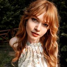 Saçları dalgalı ve omuzlarına kadar uzanıyor, kızıl saçlı, doğrudan kameraya bakıyor. Üzerinde çiçek desenli, açık renkli bir elbise var. Arka planda yeşil yapraklı ağaçlar ve ahşap bir çit görülüyor. Yüzü hafif bir gülümsemeyle aydınlanmış.