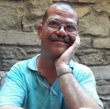 Mavi tişörtüyle, sol eli üzerine yanağını yaslamış tebessüm ediyor. Hafif dökülmüş saçları,gür bıyıkları ve gözlüğü var.