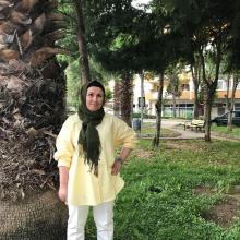 Bir palmiye ağacının önünde açık sarı gömleği, beyaz pantolonu, koyu yeşil baş örtüsüyle sol eli belinde tebessüm ediyor.